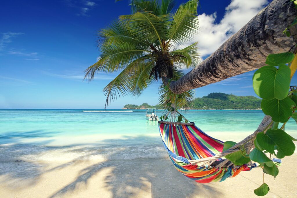 Bunte Hängematte, die an einer Palme über einem idyllischen, weißen Sandstrand hängt, mit türkisfarbenem Meer im Hintergrund. Der Blick auf das ruhige Wasser und die grünen Hügel der Küste vermittelt eine Atmosphäre von Ruhe, Entspannung und tropischer Gelassenheit.