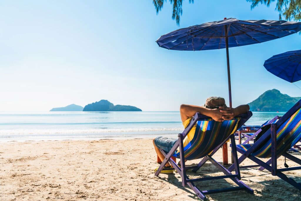 Person entspannt sich auf einem Liegestuhl am sonnigen Strand, mit Blick auf das ruhige Meer und eine entfernte Insel im Hintergrund. Ein Sonnenschirm bietet Schatten, während die Szene eine Atmosphäre von Ruhe und Erholung vermittelt.