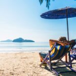 Person entspannt sich auf einem Liegestuhl am sonnigen Strand, mit Blick auf das ruhige Meer und eine entfernte Insel im Hintergrund. Ein Sonnenschirm bietet Schatten, während die Szene eine Atmosphäre von Ruhe und Erholung vermittelt.