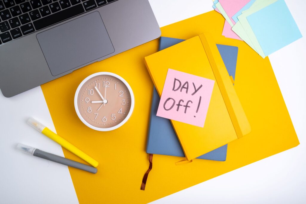 Schreibtischszene mit einem Laptop, einer Uhr und einem Notizbuch, auf dem ein Post-it mit der Aufschrift "Day Off!" klebt. Bunte Notizzettel und Stifte liegen auf einer gelben Schreibtischunterlage, die eine bevorstehende Auszeit oder einen freien Tag symbolisieren.